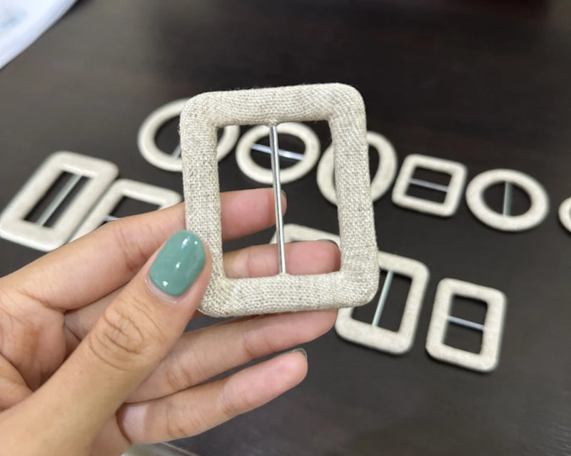 Hand holding a square fabric buckle with more buckles in the background on a dark surface